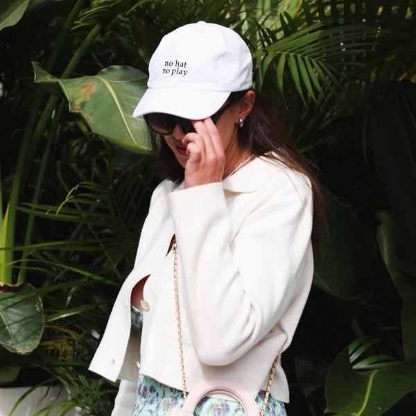 A woman wearing a white baseball cap, white jacket, and a light blue and purple floral mini skirt, carrying a light pink quilted handbag with a gold clasp, set against a background of green tropical plants.