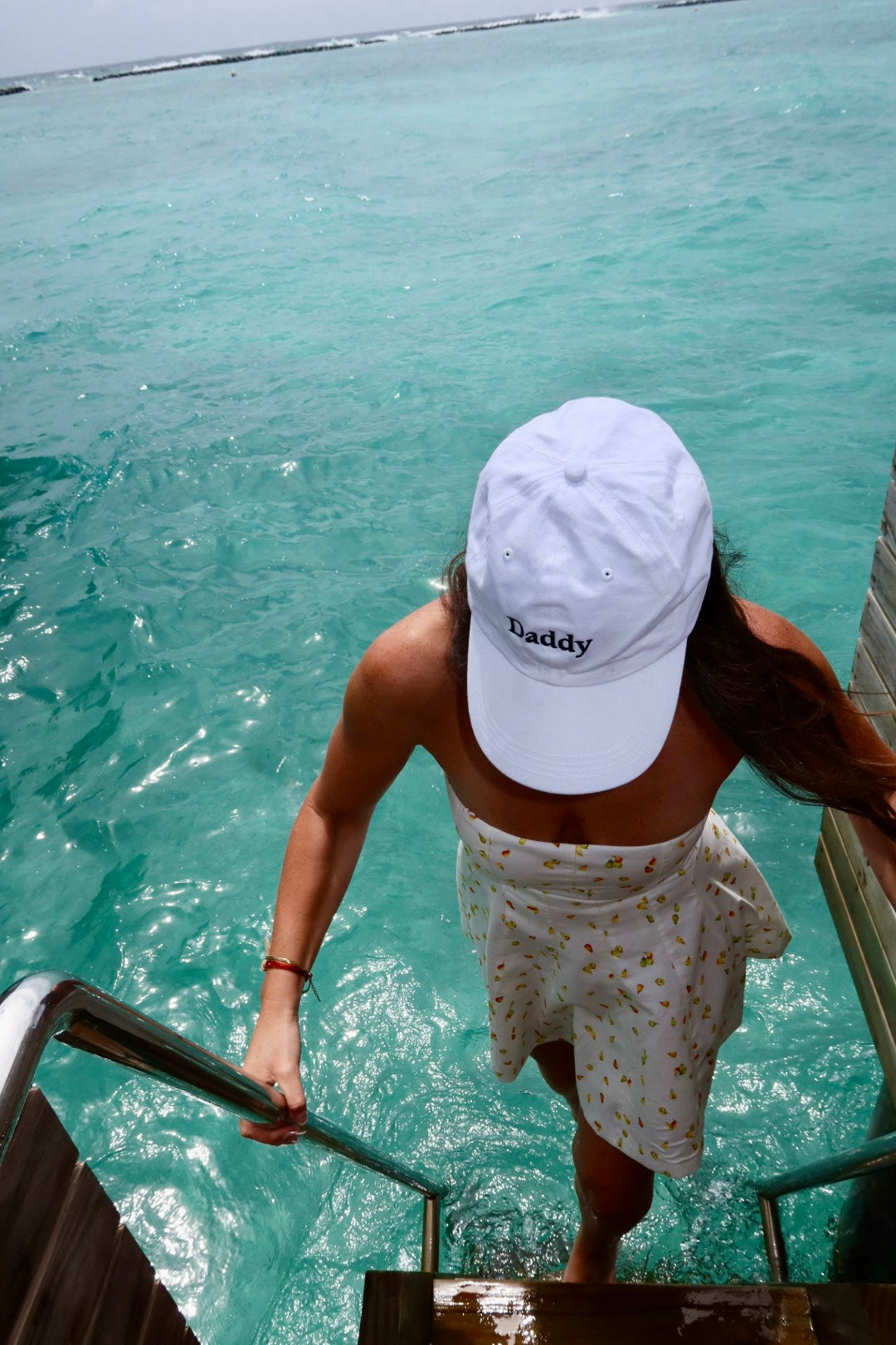 Back view of a woman wearing the white Daddy embroidered cap climbing steps from turquoise water - No Hat No Play
