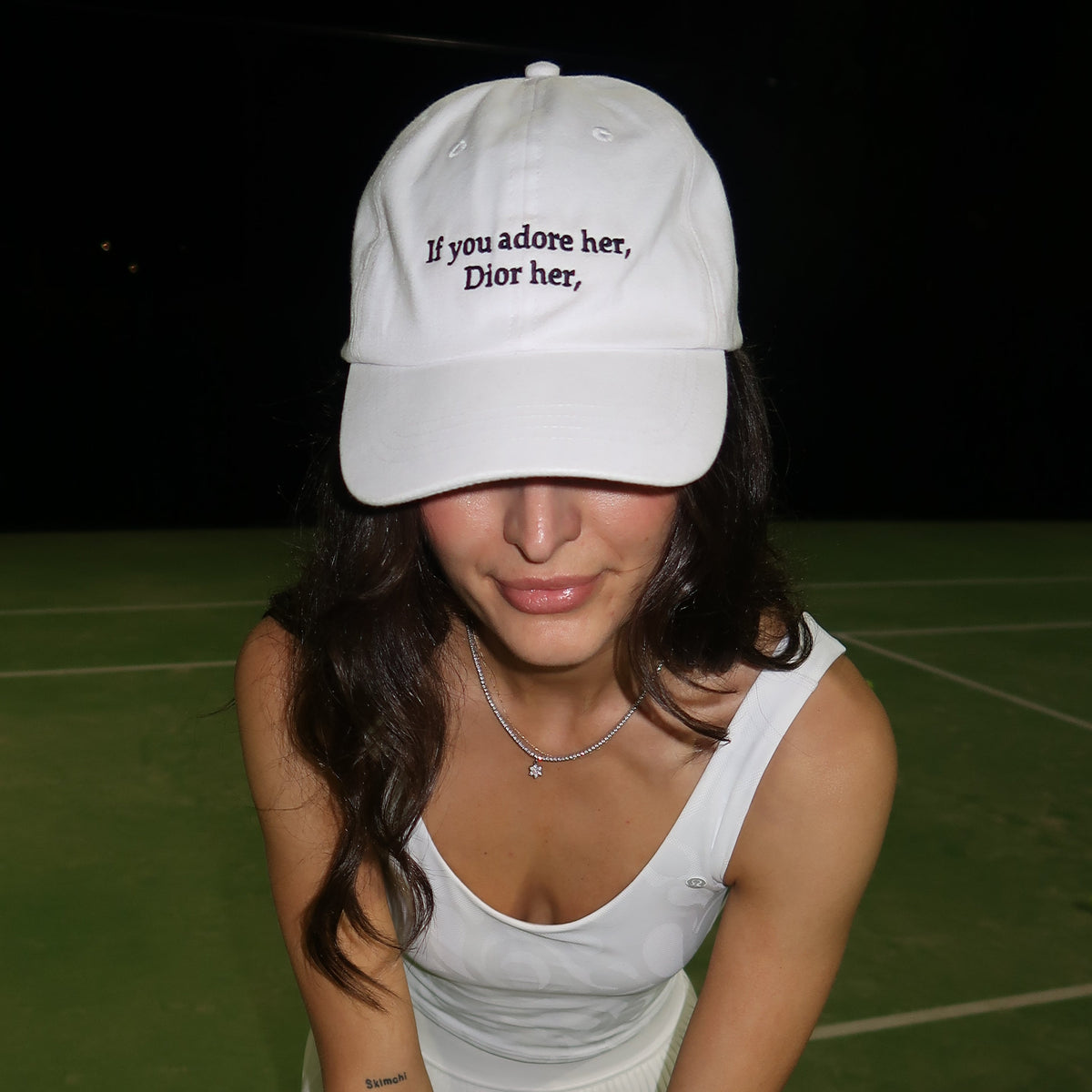 A woman on a tennis court wearing a white sleeveless top and skirt, a white baseball cap with black text, and holding a tennis racket and tennis ball.