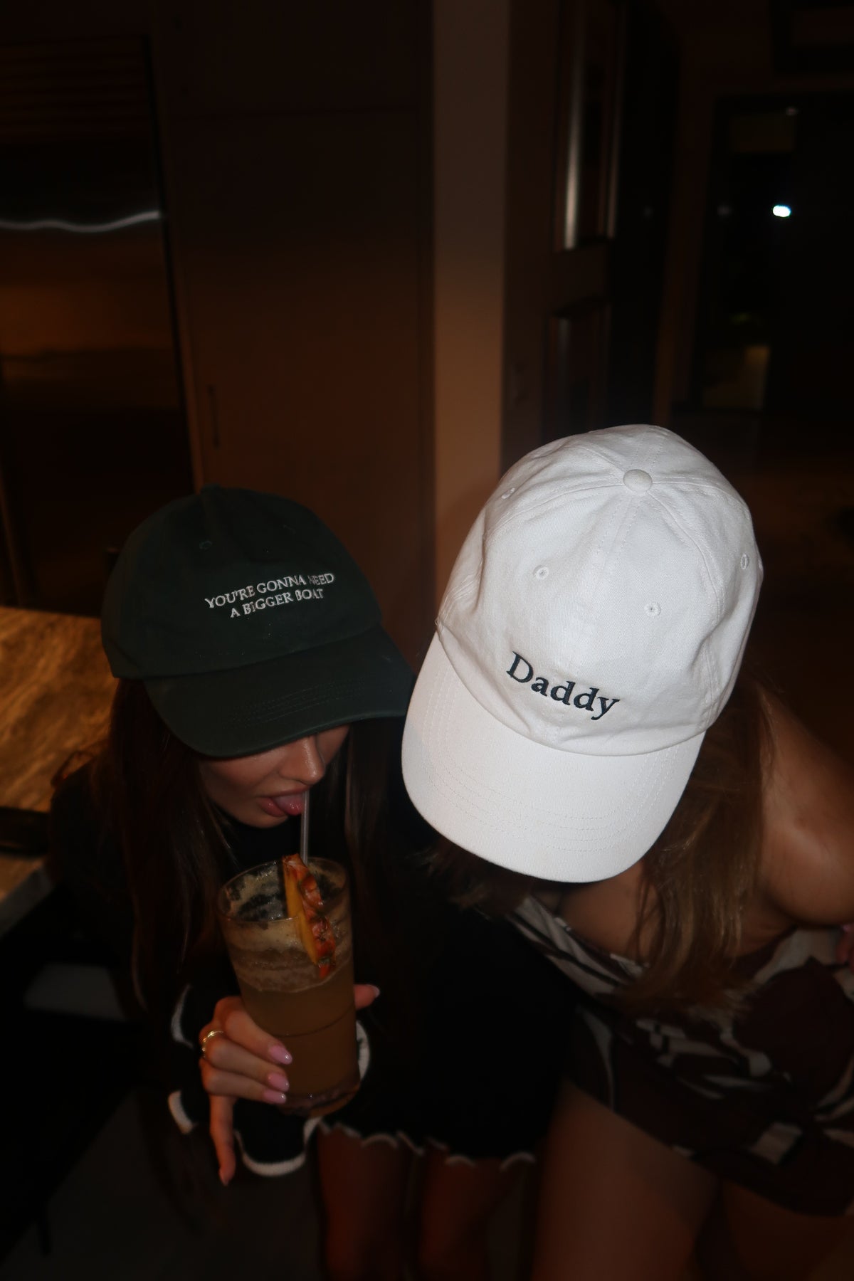 A person in a dimly lit indoor setting wearing a white baseball cap with the word 'Daddy' embroidered in black on the front, and another person wearing a dark green cap holding a drink.