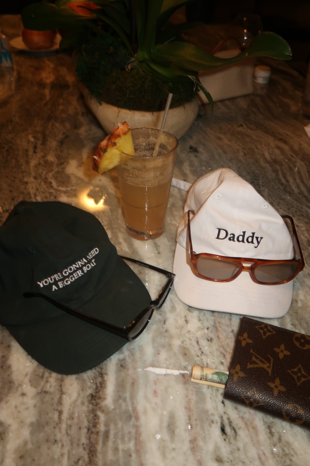 A styled flat-lay featuring a white baseball cap with 'Daddy' printed on it, a black cap with white text, sunglasses, a Louis Vuitton wallet, and a drink on a marble surface.